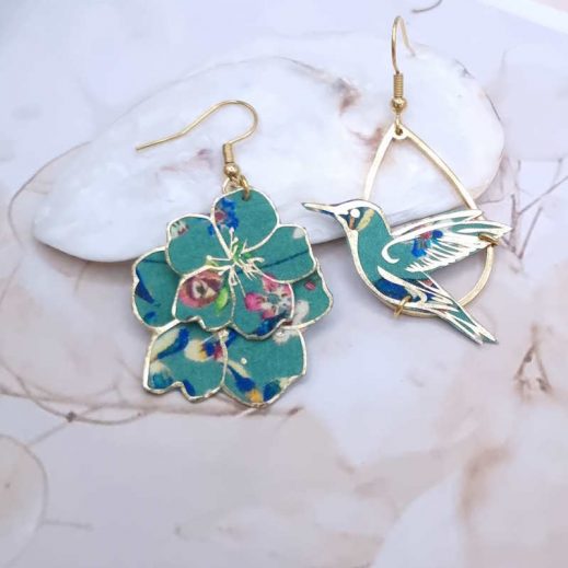 Apportez une touche de poésie et d'originalité à votre style avec ces boucles d'oreilles asymétriques uniques. Elles présentent un colibri prêt à s'envoler d'un côté et une délicate cascade de fleurs de l'autre, toutes deux réalisées à partir d'un tissu précieux vert pétrole aux motifs floraux et dorés. Faites main en France, légères et dorées à l'or fin, elles mesurent environ 4 cm de long. Parfaites pour celles qui osent un bijou distinctif et plein de charme.