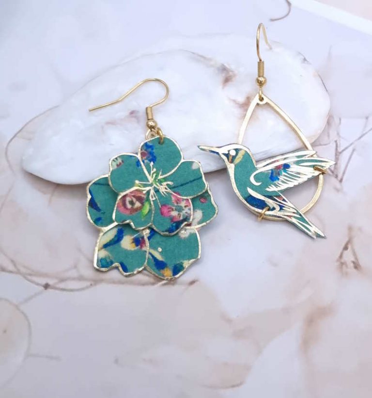 Apportez une touche de poésie et d'originalité à votre style avec ces boucles d'oreilles asymétriques uniques. Elles présentent un colibri prêt à s'envoler d'un côté et une délicate cascade de fleurs de l'autre, toutes deux réalisées à partir d'un tissu précieux vert pétrole aux motifs floraux et dorés. Faites main en France, légères et dorées à l'or fin, elles mesurent environ 4 cm de long. Parfaites pour celles qui osent un bijou distinctif et plein de charme.